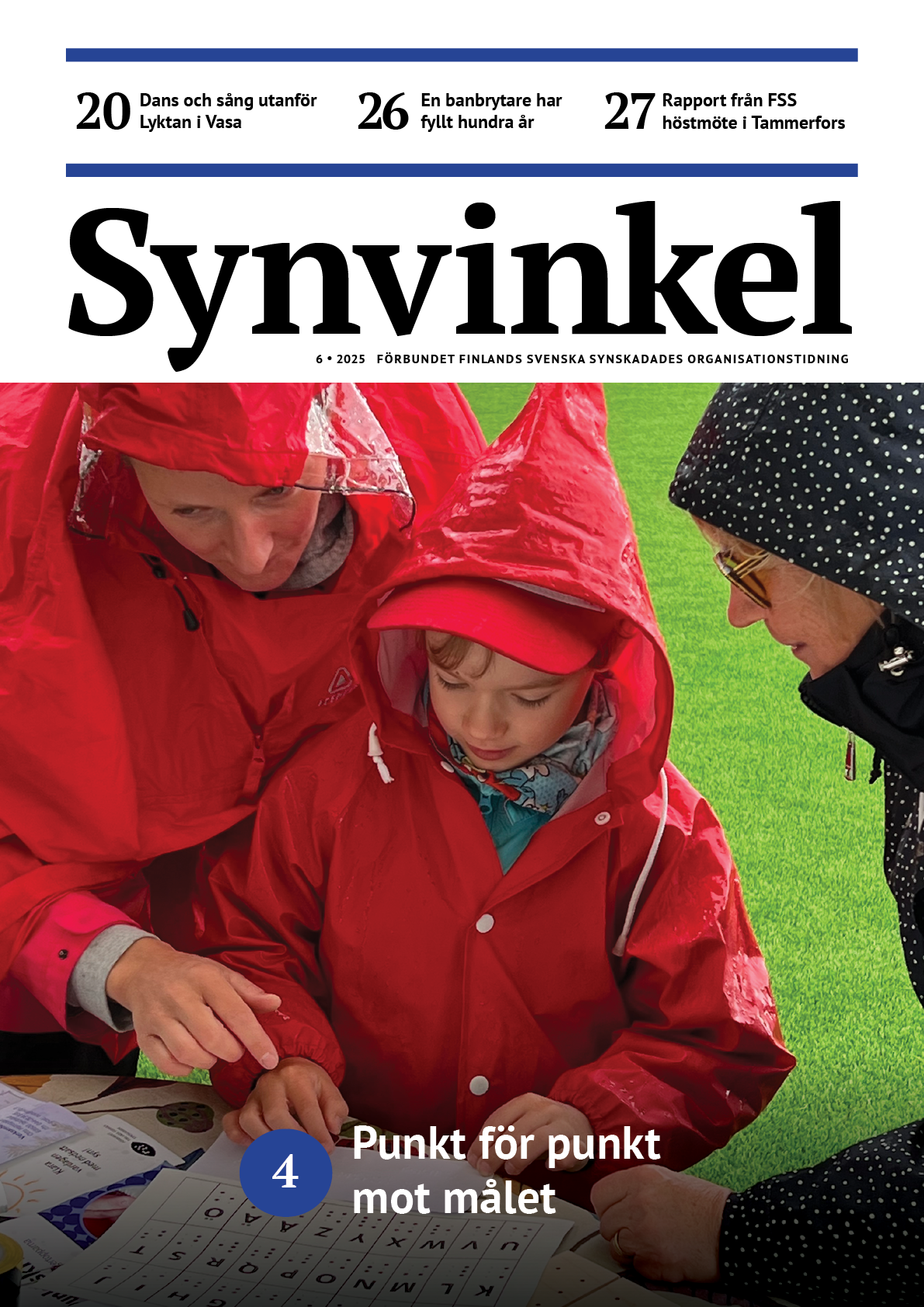 En kvinna och ett barn klädda i röda regnrockar med huva studerar en lapp i punktskrift som ligger intill ett punktskriftsalfabet på ett bord på en sportplan med grönt gräs. En kvinna i svart regnrock med vita prickar ser på.