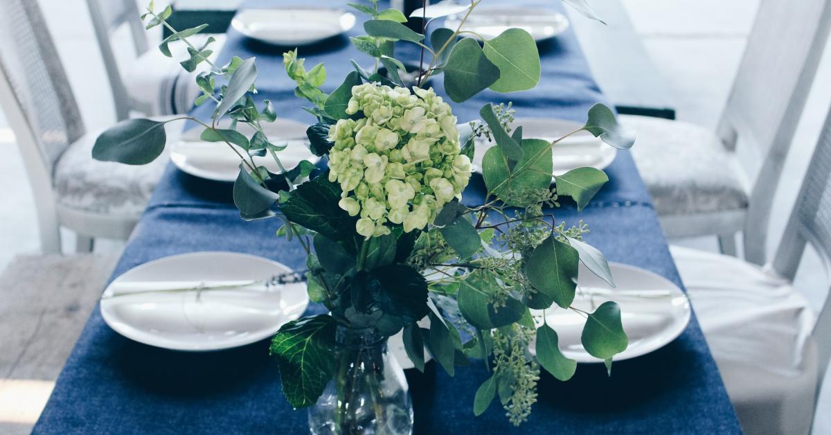 Ett långbord med mörkblå duk och vita tallrikat. Blommor i en vas på bordet. Ljusa stolar.