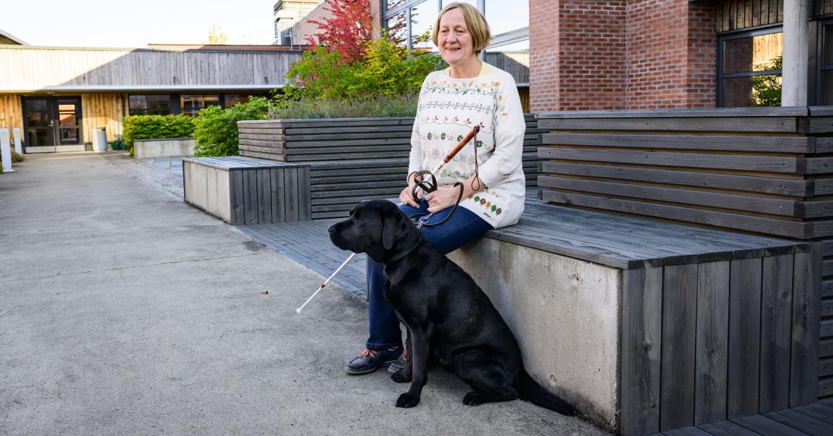En kvinna med vit käpp sitter på en bänk. Hon håller i kopplet till en svart labrador. Kvinnan har pagefrisyr och en gräddvit tröja med mönster av blommor och blad i grönt, brunt och gult.