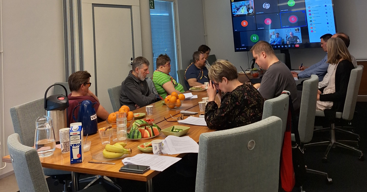 10 personer sitter kring ett mötesbord. På en stor skärm ser man att ett distansmöte pågår med 8 deltagare/deltagargrupper.