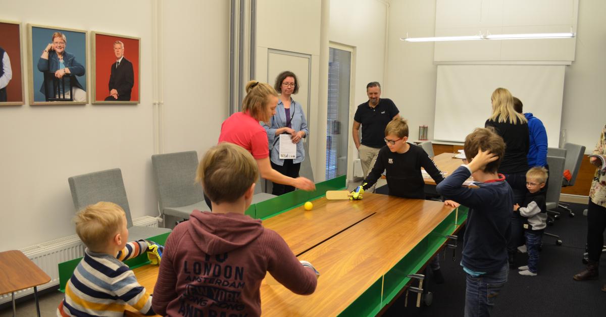 Barn och fullvuxna runt ett bord där det spelas bollspel med klubba.