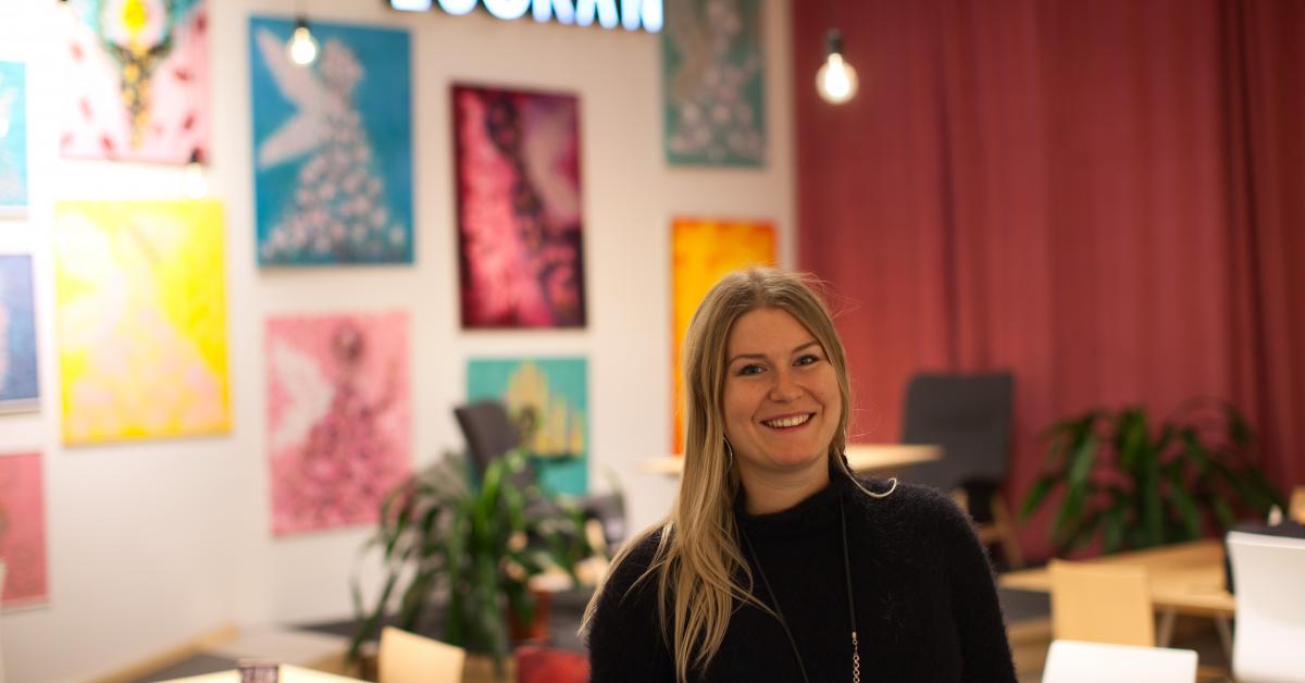 Elin Sundell med långt blont hår och svart tröja ler mot kameran. Bakom henne ett rött draperi och tavlor på väggen och en Luckan-skylt.