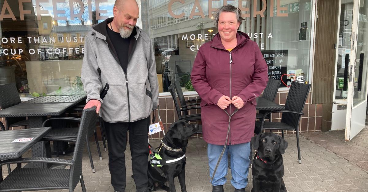 Anders med vitt skägg och grå jacka och skrattande Jessica i vinröd jacka står framför café med två svarta labradorer varav den ena har sele och den andra koppel.