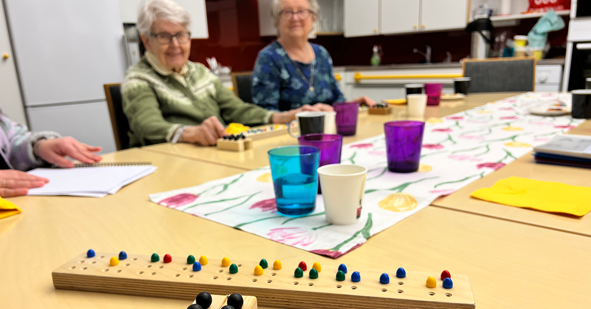 Ett bord med granna glas. I förgrunden händer intill ett ordbräde med granna knappar för undervisning i punktskrift. I bakgrunden två glada damer med var sitt bräde.