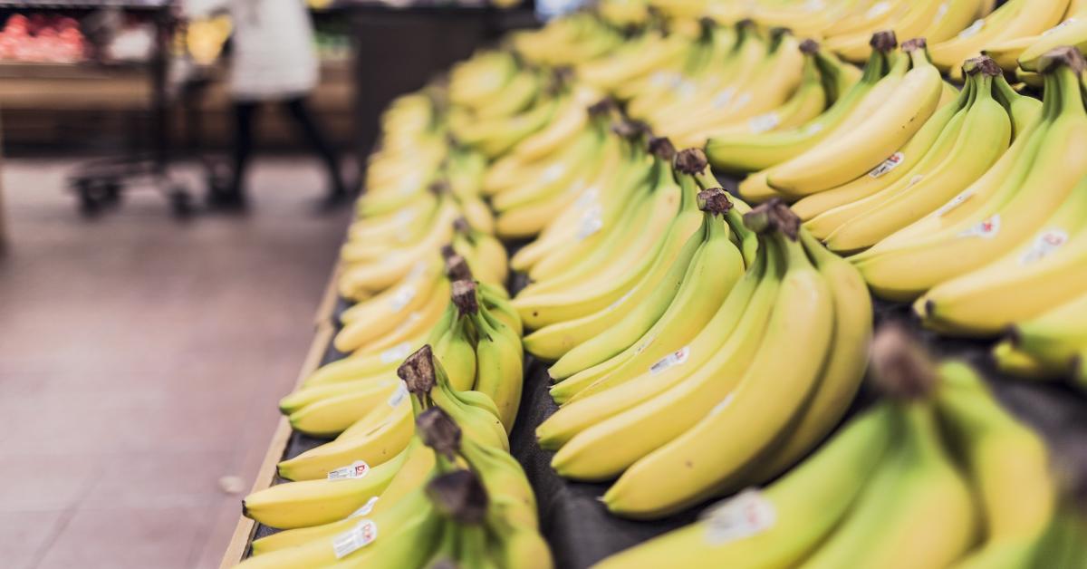 En butishylla fylld med färska bananer. I bakgrunden syns en kvinna som går med butikskärra förbi en kallhylla.