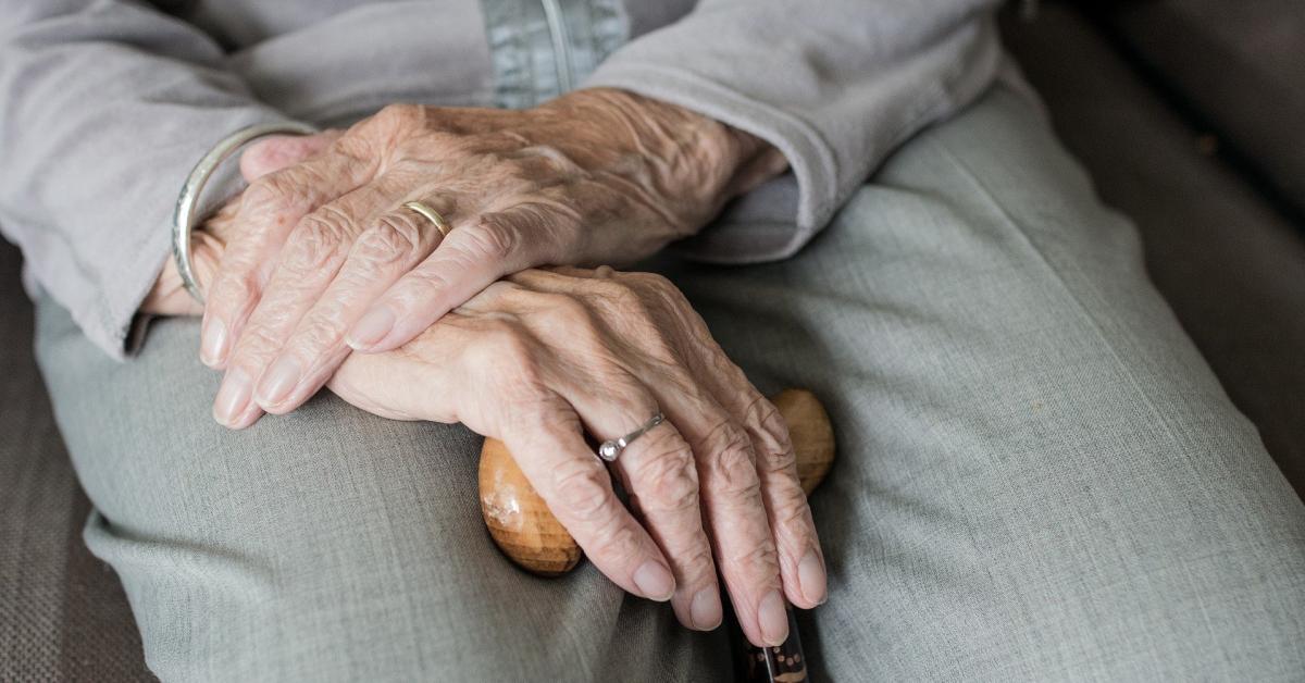 En bild på en gammal kvinnas händer. Hon sitter ner och håller i en käpp. 