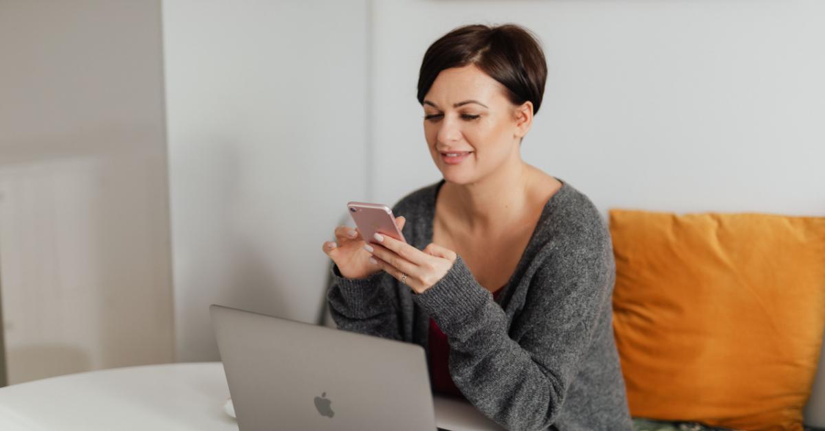 Brunett medelålderskvinna sitter på soffa vid bord med laptop, ler och skriver på smarttelefonen.