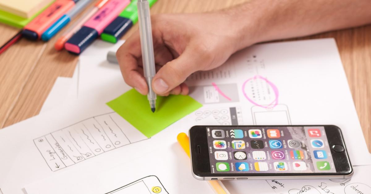 En mans hand skriver med penna på post-it lapp. Bredvid ligger en smarttelefon, tusher i olika färger och papper på ett bord.