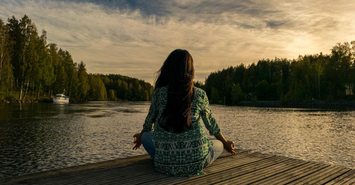 Långhårig kvinna sitter på brygga vid sjö och mediterar.