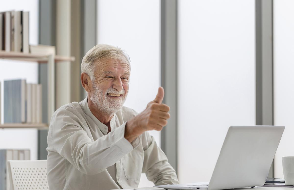 En brett leende äldre vithårig herre med skägg sitter vid en bärbar dator i ett ljust arbetsrum och håller upp tummen. 