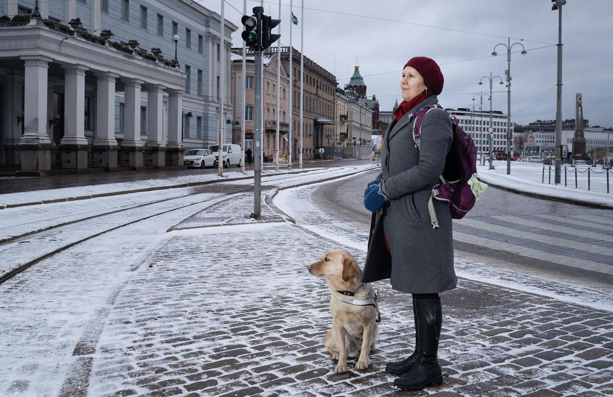 Veera Florica Rajala står med ledarhunden Palo vid en övergång på Salutorget i Helsingfors i vintrigt landskap. Tillsammans med FSS-distrikten och övriga FSS-anställda jobbar hon med påverkan av makthavarna i den nya riksdagen, i städerna och i vårddistrikten.