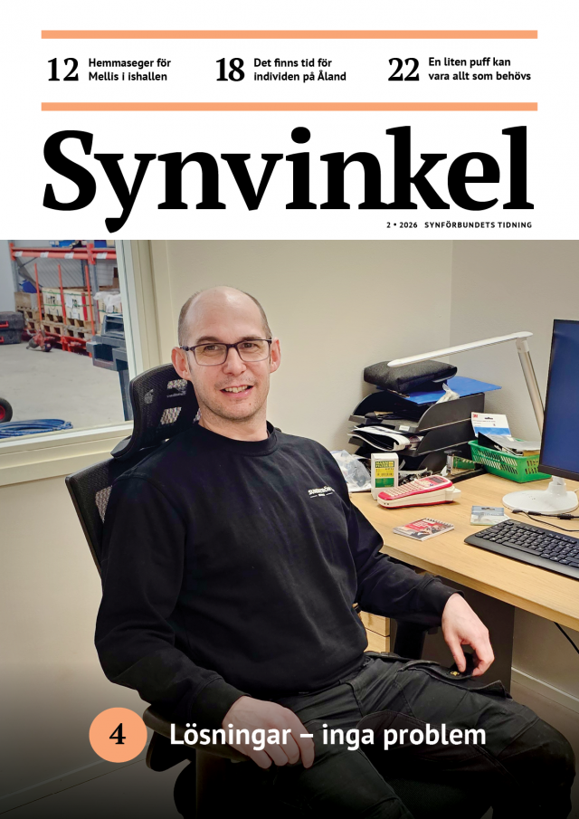 En leende, medelålders man med glasögon sitter i en kontorsstol vid ett skrivbord. Han är klädd i svarta arbetsbyxor och en svart långärmad tröja med texten Sundström på bröstet. Genom ett fönster bakom honom syns ett lager med röda hyllor.