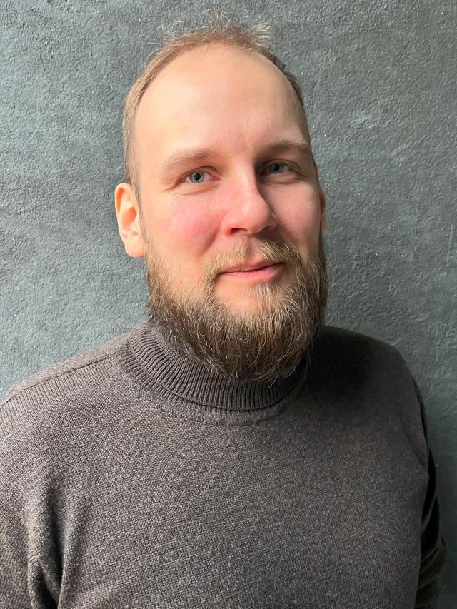 Andreas har blå ögon, brunt hår, högt hårfäste, skägg och mustasch och är klädd i en grå tröja.