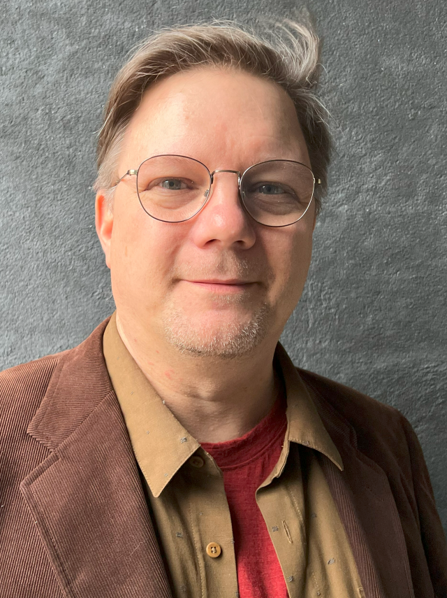 Christian har mellanljust kort hår, ovala glasögon och brun kavaj. Han ler.