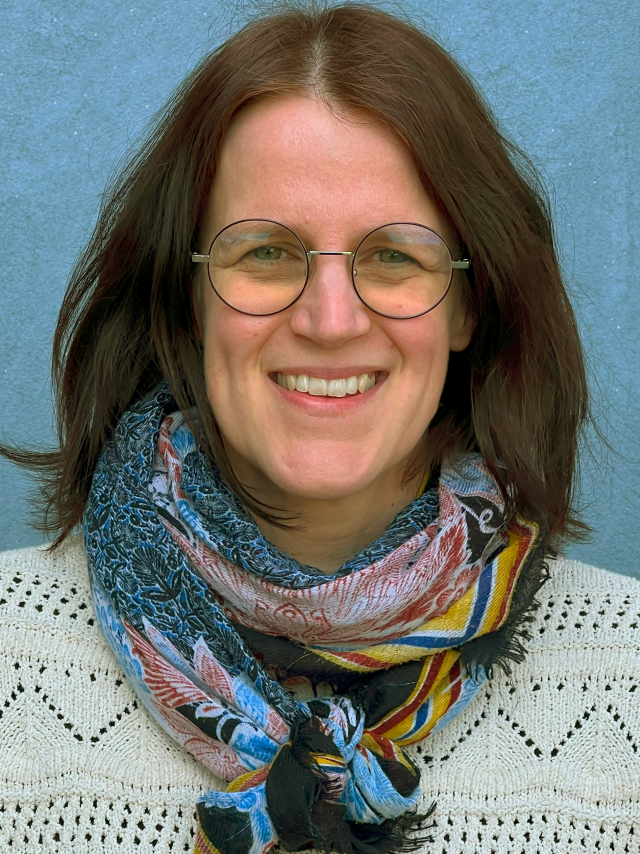 Halvfigur av leende kvinna med mörkbrunt, axellångt hår, runda glasögon med tunna bågar. Hon är klädd i en naturvit mönsterstickad tröja och en mönstrad scarf. 