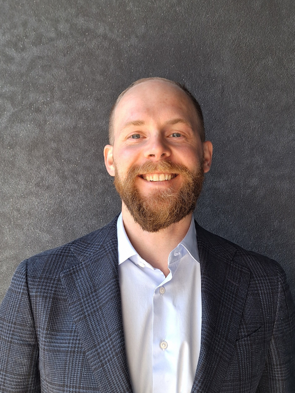 Anton har blå ögon, brunt hår, högt hårfäste, skägg och mustasch. Han är klädd i kavaj